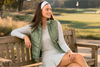 Female golfer sitting on a bench wearing a sage green winter golf vest