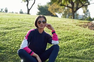 Woman resting on the golf green in her Navy, Blossom, White accent sweater and navy pants. Sunshine behind her she is happy.